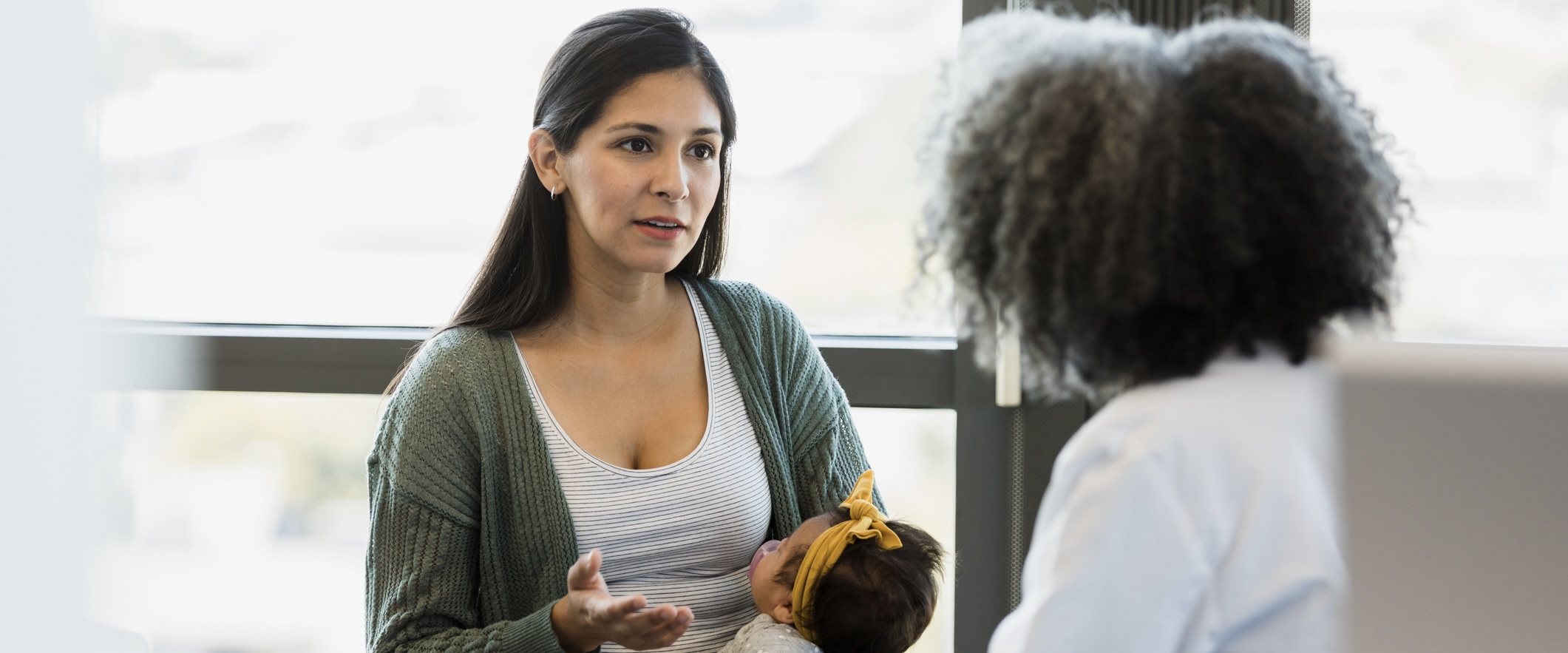 Woman gestures while holding a baby and speaking with doctor.